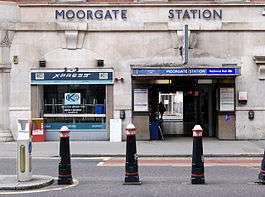 A grey building with a rectangular, dark blue sign reading "MOORGATE STATION" in white letters and short, black, white-tipped posts in front