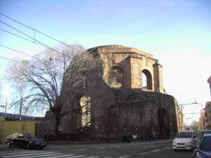 Exterior of a ten-sided ruin called today the Temple of Minerva Medica at the intersection of city streets in Rome showing large arched windows in the drum between engaged buttresses and below polygonal step-rings buttresses for the collapsed dome