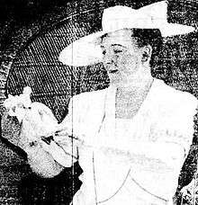 Black and white portrait of Ethel Kelly in a white dress and white hat, holding a doll
