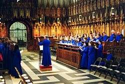 A view in the choir of York Minster with girl choristers and male lay clerks in blue cassocks standing in the richly carved choir stalls.