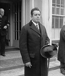 A black and white portrait of a formally dressed man standing outside a building. He is wearing a dark coat, white shirt, dark tie and he is holding a hat with his left hand.