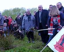 A group of people stand in a grassy field in daylight watching a woman digging the ground using a spade. People are taking pictures of the event and have brought banners and football scarves.