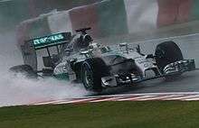 Silver Formula One car in the rain; standing water on the track's surface is lifted by its tyres.