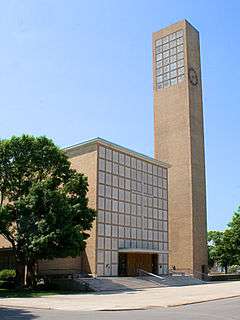 Rectangualr building with large cross and many square on front next to a tall clock tower