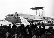 Black-and-white photograph with angled front view of four-engine jet aircraft on ramp with front fuselage door opened: A contingent of people are there to welcome the jet, which has a disc-shaped radar perching on top of struts on the dorsal fuselage.