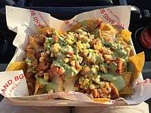 A cardboard tray lined in white paper with "The Band Box - Right Field" holds yellow corn chip, smoked chicken pieces, jalapeño queso, sweet yellow corn, pico de gallo, and green cilantro crema