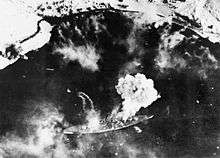 Black and white photograph of a warship moored near a snowy shore viewed from the air. Smoke is issuing from the warship.