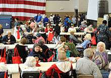 Image of a large number of primarily elderly people sitting at long tables within a building, with the United States flag in the background.