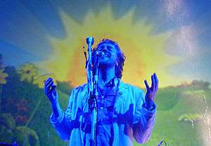 A man standing behind a microphone stand; a blue light is shining on him and a colorful, picturesque effect appears in the background.