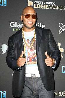 An Afro-American male donning a black peacoat, ash-colored jeans, dark sunglasses, a white shirt with scarlet lettering, and a gold chain about the throat, gives the thumbs up gesture on back hands while smiling in front of a gray background
