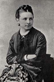 A woman with tight hair and a dark shawl top sits with her arms crossed, facing to the viewer's right with her body turned to the viewer's left