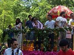 A parade float with fencing is decorated with flowers. Multiple men and women of South Asian descent lean towards fans on the street.