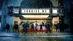 A faraway image of a woman and many men. She wears a pink raincoat with black leggings. The men wear black clothes and each carry an umbrella. Behind them can be seen a theatre, on the marquee is written "Forgive Me" in black capital letters. Also, it is raining.