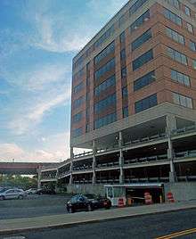 A parking lot next to a multistory parking deck, with a tall modern concrete, brick and glass high-rise at right