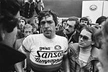 A man sweating and wearing a cycling jersey while being surrounded by several people.