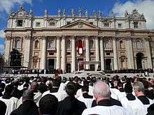  The exterior of the basilica on a sunny day. In the foreground, hundreds of robed priests look towards a podium where there is an altar, and a group of white robed figures attends the Pope.