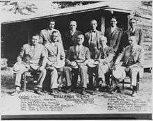 Black and white photograph of several political figures seated in front of a rustic building, FDR in the center of the group.