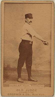 A baseball player is shown standing in his uniform, holding a baseball with his arm stretched out across his upper body.