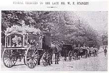 Black and white press photograph showing the funeral procession. The title is "Floral tributes to the late Mr. W. F. Stanley". There are four vehicles shown. From left to right: The hearse horse and carriage, covered in flowers, with an undertaker climbing up onto the carriage. In front of the hearse, there are a horse and carriage, carrying mourners. In front of this carriage are two further horses-and-carriages carrying flowers.