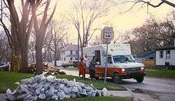 Several people are collecting items from an American Red Cross vehicle in a neighborhood. Around the neighborhood is piles of damaged possessions and sandbags.