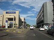 A road with a Bank Gaborone building to the left and a taller building with curved truss structures on the windows to the right