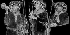 A black and white image of three aged men wearing hats. On the left a man playing the gaita, another man at the centre singing to a microphone and on the right another man playing the gaita.