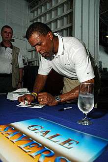 A man looks down as he signs autographs for fans on a table. He is wearing a white polo shirt and khakis.