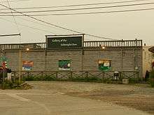 A one-storey building with a log front and part of a concrete-block side facade visible. On the log facade is white lettering spelling out "Gallery of the Midnight Sun" and paneling with large artworks depicting natural scenes