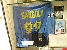 A blue coloured T-shirt displayed at a store window. The T-shirt has the words "Ganguly" and the number 99 below it, both in yellow color. Beside the T-shirt, a picture and an open book is visible.
