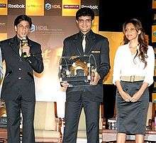 Two middle-aged males and one female standing. The man in the middle wears a black suit and carries a golden coloured casket. The other man to his right wears a black suit and speaks in a microphone. The lady on the left wears a white shirt and black skirt. Her hair is brownish and falls in locks around her.