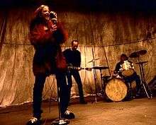 A female singer and a male bassist and drummer perform against a golden backdrop.