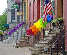 Stoops on some brick houses, seen looking along the sidewalk. One has balloons in the colors of the rainbow tied to its railing. Another has a purple ribbon.