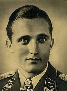 The head and shoulders of a young man, shown in semi-profile. He wears a military uniform with an Iron Cross displayed at the front of his white shirt collar. His hair appears dark, short and combed back, his nose is long and straight, and his facial expression is emotionless; looking to the left of the camera.
