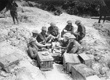 Eight men, most wearing steel helmets, sit in a shell hole surrounded by wooden crates.