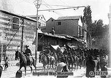 Soldiers on horseback trot along a road past several buildings watched by a group of civilians