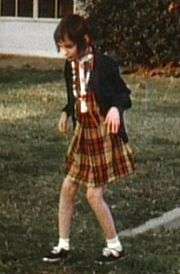 A slightly blurry color picture of Genie, facing slightly right of the camera, walking by herself outside in the Children's Hospital recreation yard. She is wearing a plaid-patterned dress and thin sweater and looks extremely pale, emaciated, and expressionless. Her limbs are exposed and look extremely thin. Both of her knees are very bent, and her arms are bent forward with both hands hanging down as she holds them out in front of her.