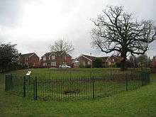 A shallow depression in the grass with a stone lined pit in the centre. It is surrounded by railings with a small interpretation board next to the gate. Behind the shallow depression is an earth bank, and behind that a row of recent houses. A large leafless tree is on the left
