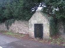 A small stone building with wrought iron gates against a long stone wall.  Huge yew trees overhang the wall, and a straggly bit of ivy clings to theside of the well house