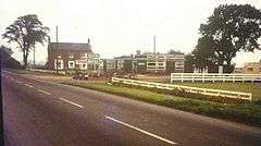 A faded period photograph of the motel on a grey day.  The main road is in the foeground, from lower right to half way up the left side.  In the middle distance a two-story 18th or 19th century building stands at right angles to the road, it's windowed frontage facing our vantage point.  It is on the opposite side of the road to us, beyond the entrance track.   This side of the track, in the centre of the photograph and still on the opposite side of the road to us are a range of low, flat-roofed, single story buildings, almost certainly new at the time.  There is low whir post-and-rail fencing on the verge this side of the motel.  The older building is probably red brick. The new buildings have dark grey walls.  The sign reading 'Trenton Motel' is clearly visible by one of the modern buildings.