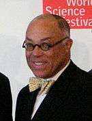 An African American male with graying hair and round eyeglasses, wearing a suit with a bow tie, set against a white and red background reading World Science Festival.