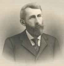 A man in his forties, facing right, with thick black hair, a beard, and a mustache. He is wearing a white shirt, black tie, and black jacket