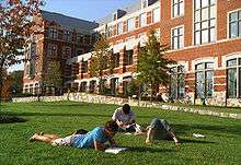 Three young adults lie on grass reading books in front of a brick building with many windows.