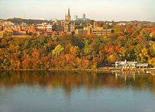 A panorama of numerous buildings, particularly the tall clocktower, above a stretch of brightly colored autumn trees all reflected in a river.
