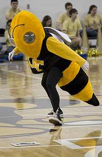 A person wearing a costume which resembles a yellowjacket, including a black shirt with yellow interlocking G-T logo, spins a dial on a wooden gymnasium floor.