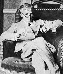 A middle-aged man sitting with his legs crossed in an armchair, looking over the top of his glasses while holding a book