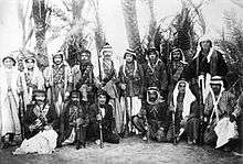 A group of rebels in traditional Arab dress posing with rifles with date palms in the background