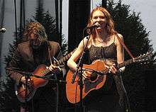 A man with his head down playing a guitar with a smiling Welch also playing guitar on stage. Both play in front of microphones.