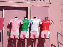 Four cycling jerseys – pink, green, white, and red, from left to right – are arranged on mannequin torsos in front of a pink wall.