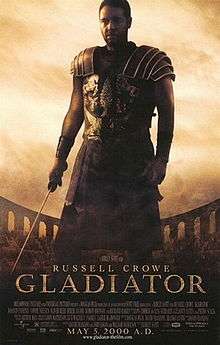 A man standing at the center of the image is wearing armor and is holding a sword in his right hand. In the background is the top of the Colosseum with a barely visible crowd standing in it. The poster includes the film's title, cast credits and release date.