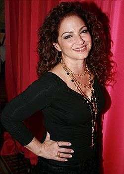 A woman with curly brown hair is standing in front of a red curtain. She is sporting a black dress and a bunch of golden chains are hanging from her neck. Her hand is placed on her waist.